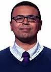 Portrait of a bespectacled man in a striped shirt, purple tie and navy sweater against white.